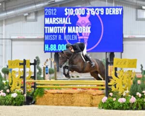 A rider in a black jacket jumps a horse over a yellow obstacle in an indoor arena, with a large scoreboard displaying event and competitor information in the background.