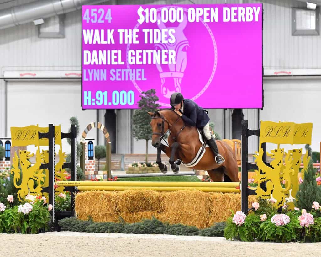 A rider on a brown horse jumps over a yellow fence during an indoor equestrian event, with a large event scoreboard in the background displaying competition details.