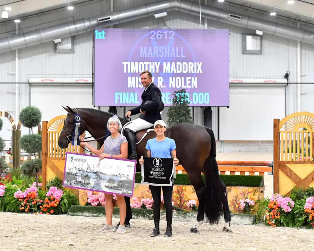 A rider on a horse poses indoors with two women holding a large check and a sign; a scoreboard in the background displays event results.