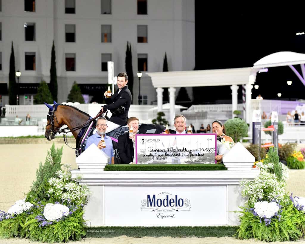 A group of people and a rider on horseback pose at an award ceremony, holding a large check and trophies, in front of a Modelo sign and floral arrangements.