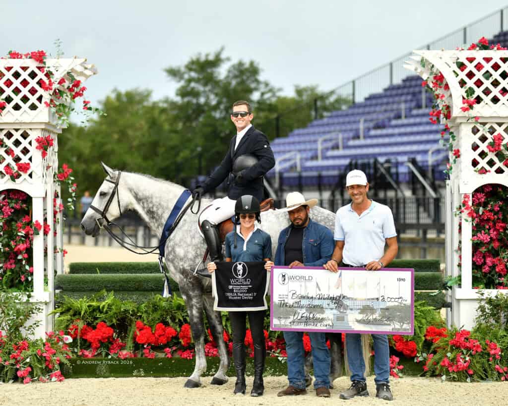 A rider on a gray horse poses with three people; they hold a trophy plaque and an oversized check in front of red flowers and a decorated arena backdrop.