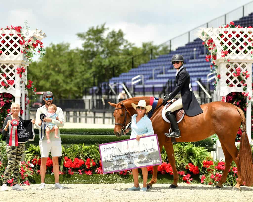 Four people and a horse at an equestrian event; one person on horseback, two holding a large check, and one holding a baby and a ribbon, with flowers and stands in the background.