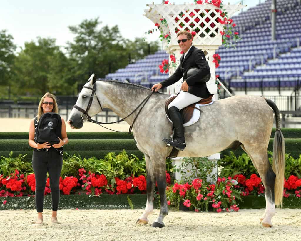 A man in riding attire sits on a gray horse next to a smiling woman holding a plaque, with flowers and stadium seats in the background.