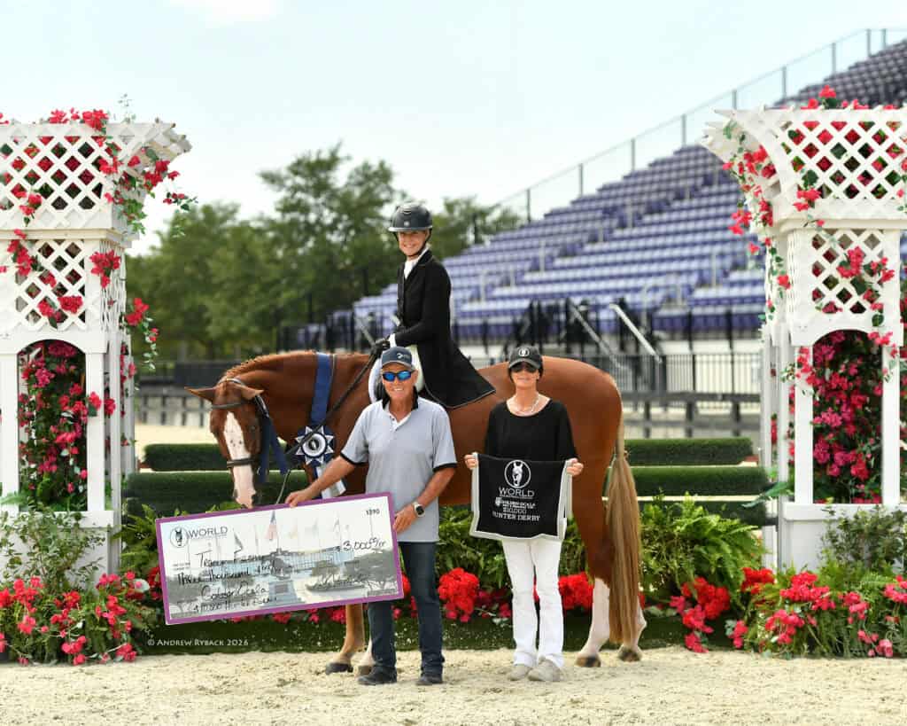 A rider on a horse poses with two people holding a large check and a trophy banner in a decorated outdoor arena.