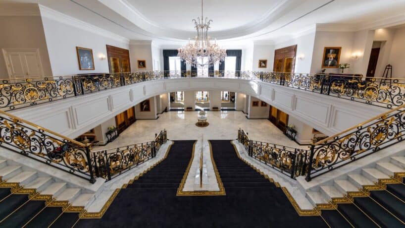 Grand staircase with ornate railings, black and gold carpet, marble floors, and a large chandelier in an elegant lobby with framed artwork and windows.