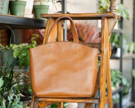 A brown leather tote bag is displayed on a wooden ladder indoors, surrounded by green potted plants.
