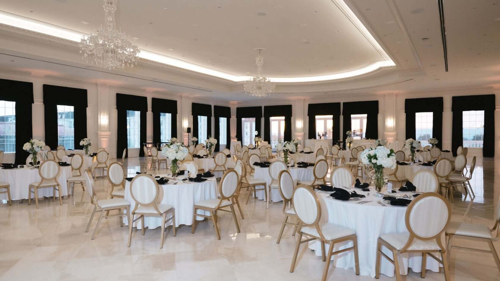 Elegant banquet hall with round tables covered in white cloths, gold chairs, black napkins, and tall floral centerpieces, with chandeliers overhead and large windows along the walls.