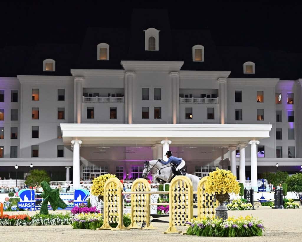 An equestrian rider jumps a horse over a yellow gate during a nighttime competition, with a large, lit building in the background and flowers in the foreground.