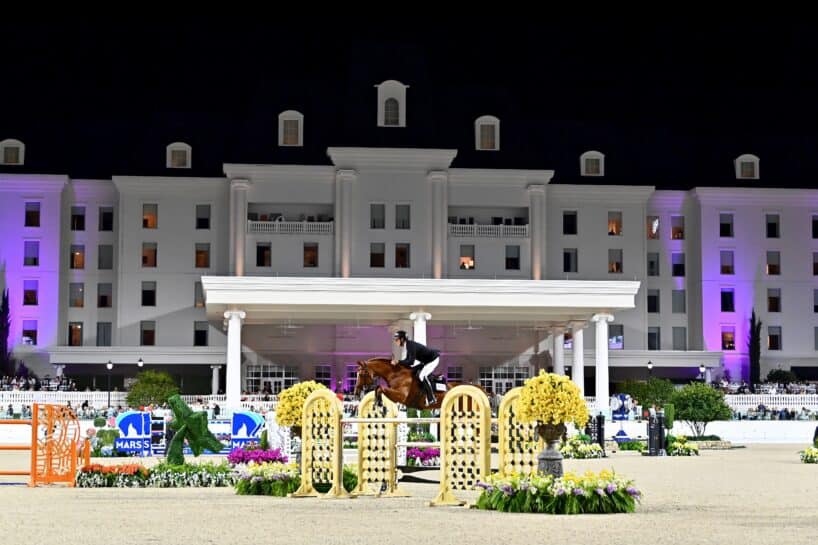 A rider on a horse jumps over an obstacle during an equestrian event at night, with a large, white building illuminated in the background.