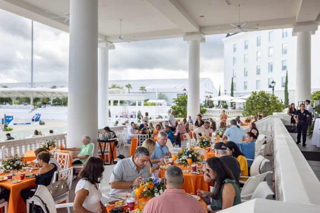 People sit at round tables with orange tablecloths on a covered outdoor patio, eating and talking. Floral centerpieces are on the tables, and a white building is visible in the background.