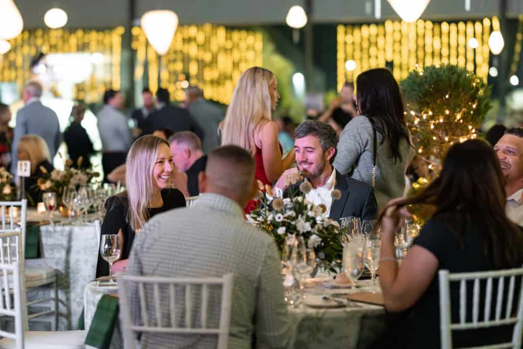 People sit and stand around decorated tables at a formal event, talking and smiling, with string lights and floral centerpieces creating a festive atmosphere.