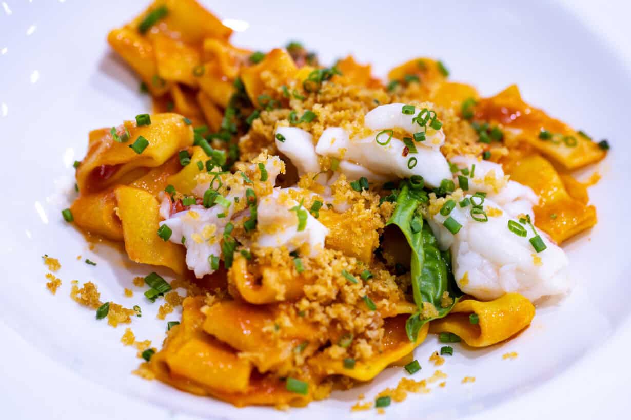 A white plate with pasta in red sauce, topped with white seafood pieces, chopped chives, green leaves, and golden breadcrumbs.