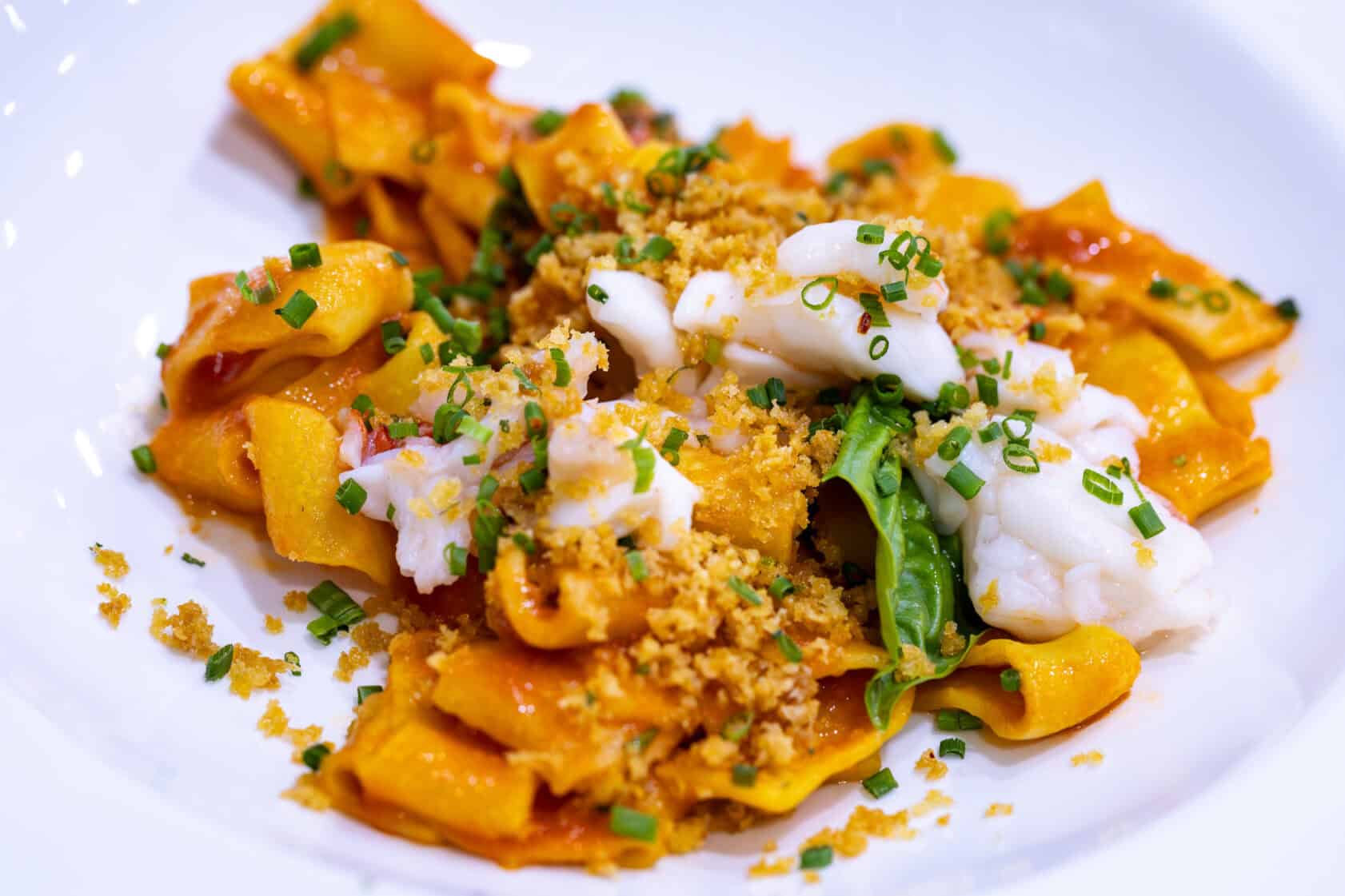 A white plate with pasta in red sauce, topped with white seafood pieces, chopped chives, green leaves, and golden breadcrumbs.