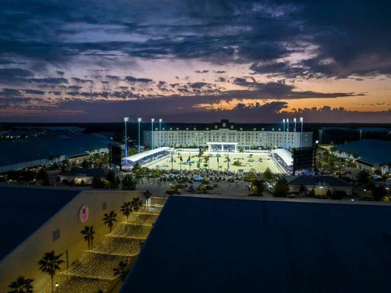 Aerial view of a brightly lit outdoor stadium hosting an event at dusk, with a large building and dramatic sunset sky in the background.