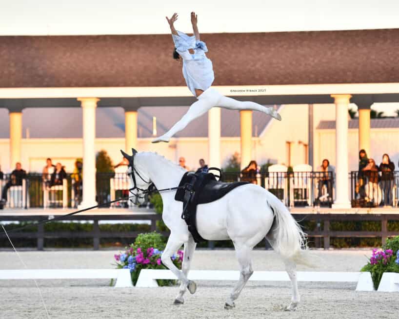 A performer in white leaps midair above a white horse during an equestrian event, with spectators and a building in the background.