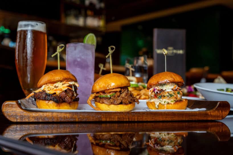 Three sliders with different toppings are arranged on a tray, with two colorful drinks and fries in the background on a restaurant table.