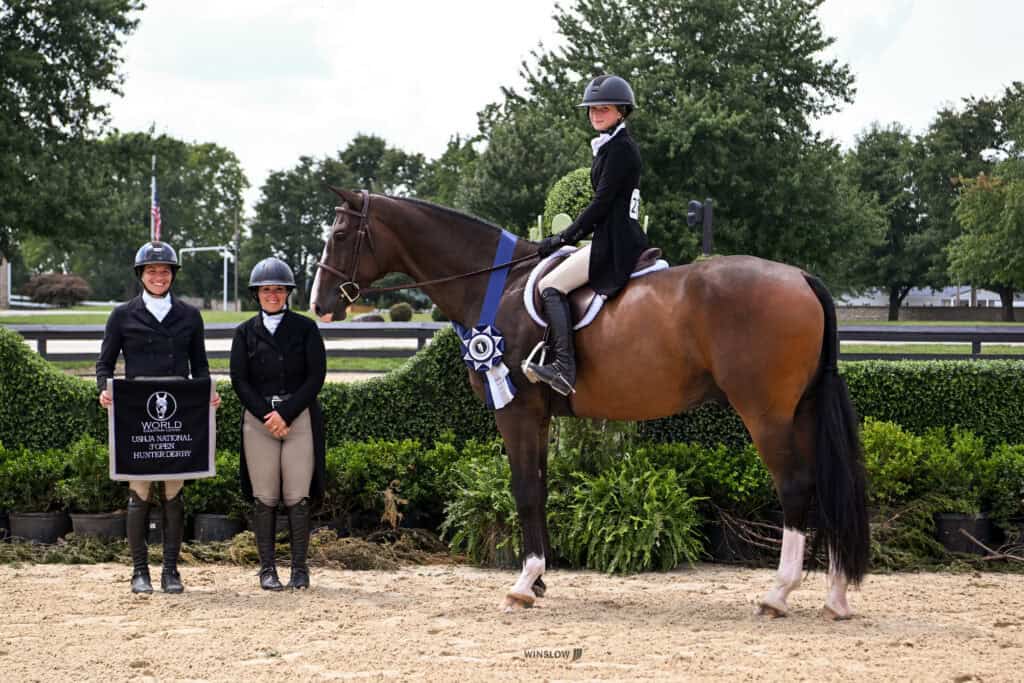 Three equestrians in riding attire pose with a horse wearing a blue ribbon. Two stand with a sign; one is mounted. Greenery and trees are in the background.