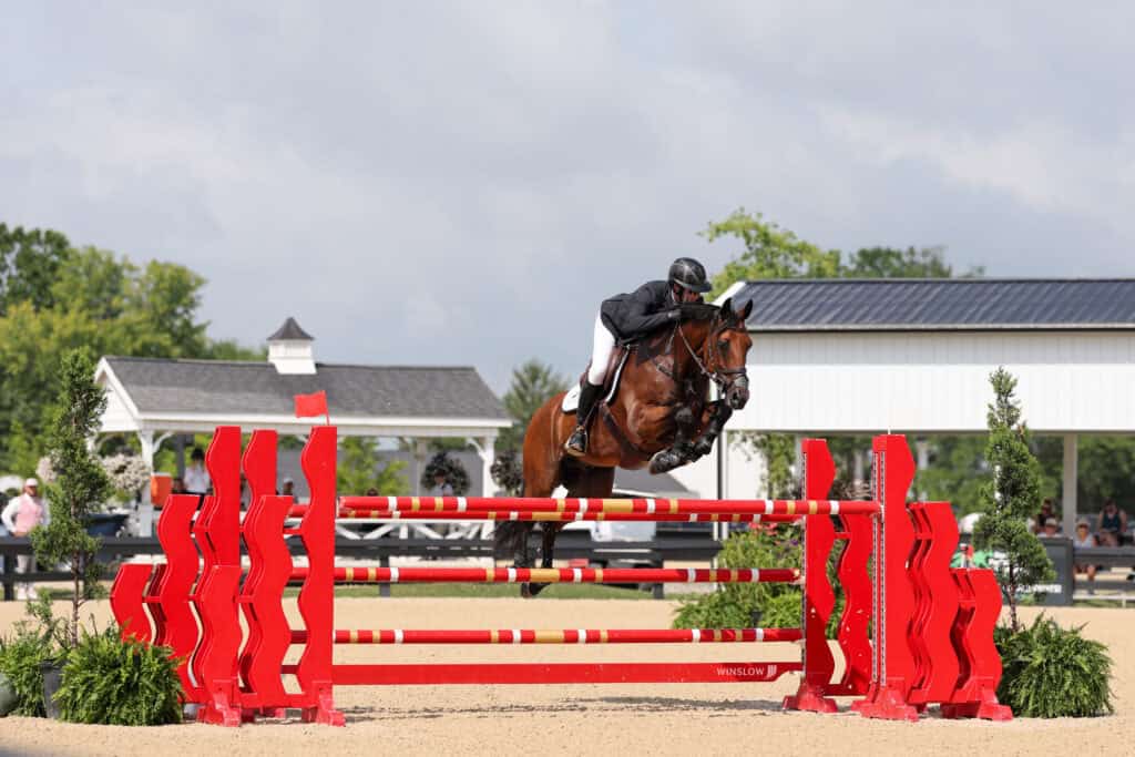 Equestrian rider and horse jump over a red obstacle during a show jumping competition at an outdoor arena.