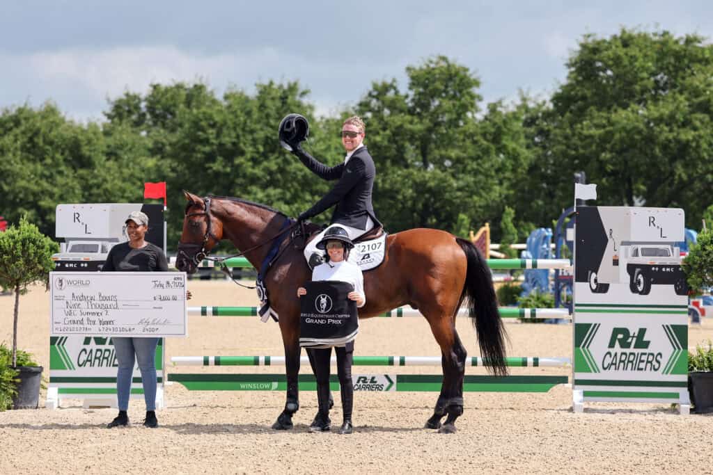 Equestrian event winner on horseback holds a helmet, flanked by two people, one holding a large check, in an outdoor arena with jumps and sponsor banners.