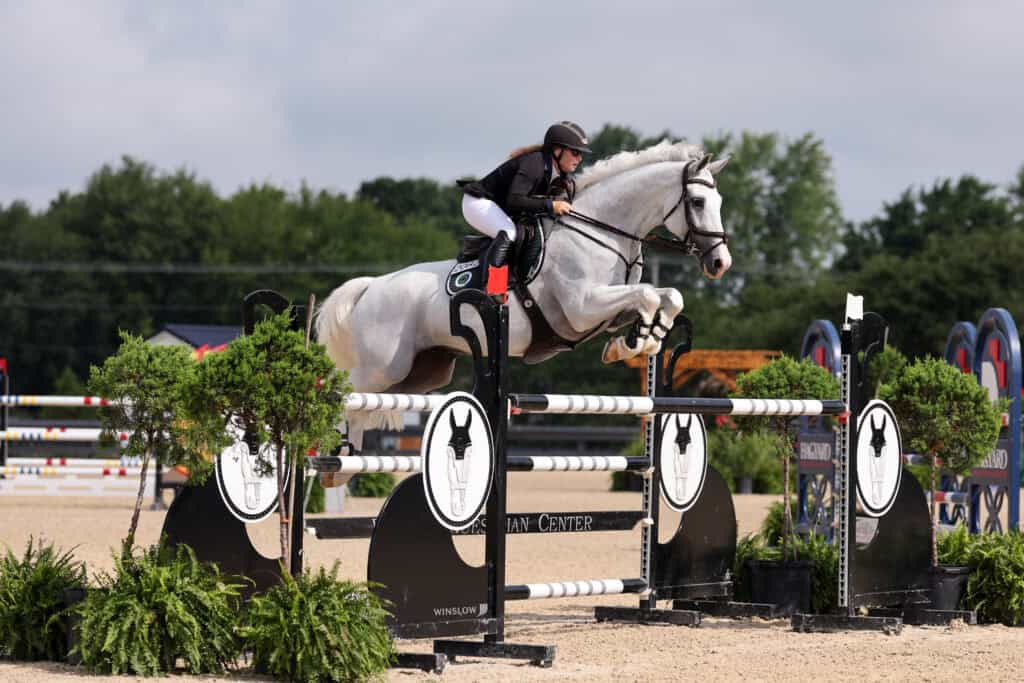 Equestrian rider in black attire jumps a gray horse over an obstacle during a show jumping competition on an outdoor sand arena.