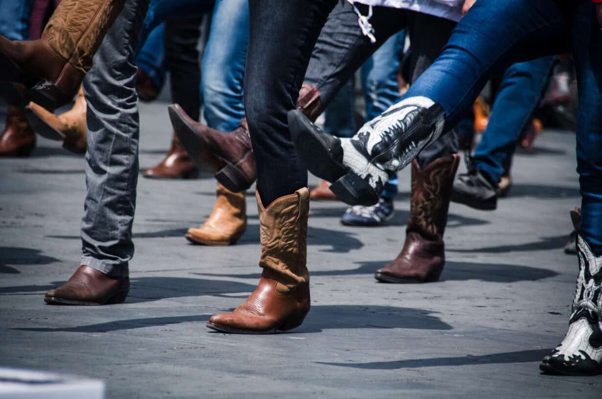 A group of people wearing cowboy boots perform a coordinated dance, lifting one leg in unison on an outdoor pavement.