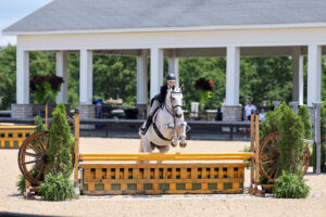 Equestrian rider in helmet and jacket jumps a white horse over a yellow and green fence at an outdoor arena.