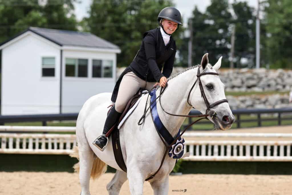 A rider wearing a helmet and jacket sits on a white horse with a blue and white ribbon, smiling at an outdoor equestrian event.