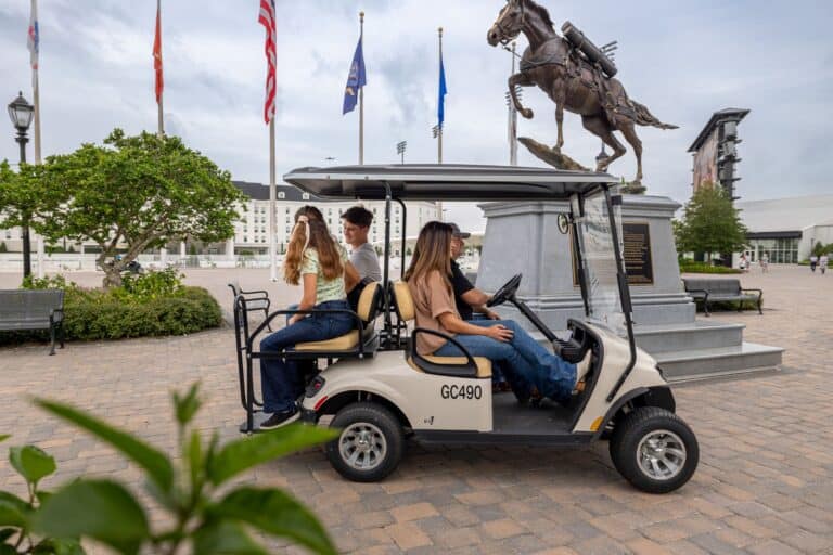 Three people ride a golf cart near a statue of a horse and rider, with flags, benches, and buildings visible in the background.