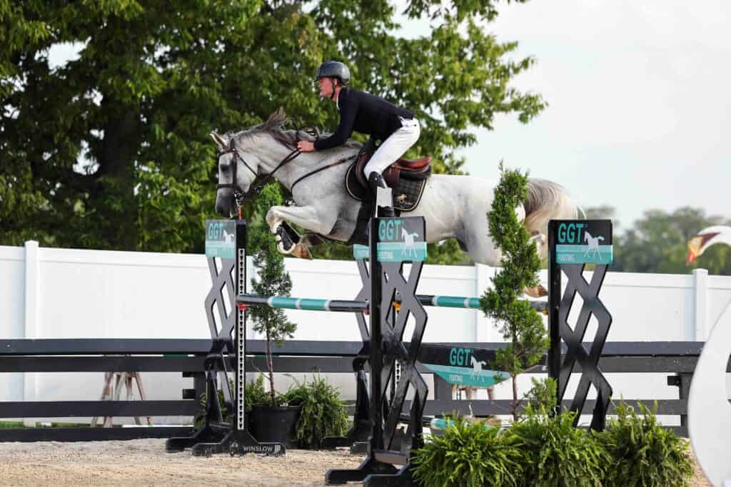 A person riding a gray horse jumps over an obstacle during an equestrian event, with trees and a white fence visible in the background.