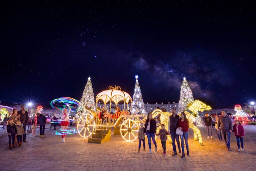 People stand near illuminated Christmas decorations including a carriage, trees, and a horse, with a night sky full of stars above them. Some children and families are posing for photos.