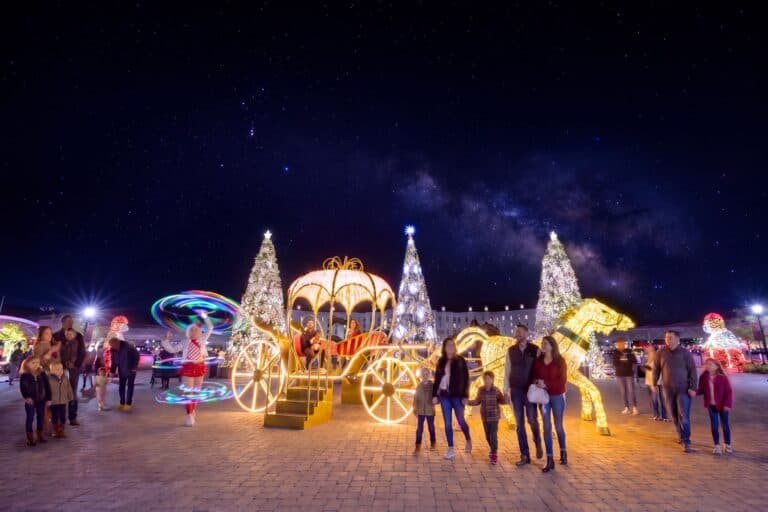 People stand near illuminated Christmas decorations including a carriage, trees, and a horse, with a night sky full of stars above them. Some children and families are posing for photos.