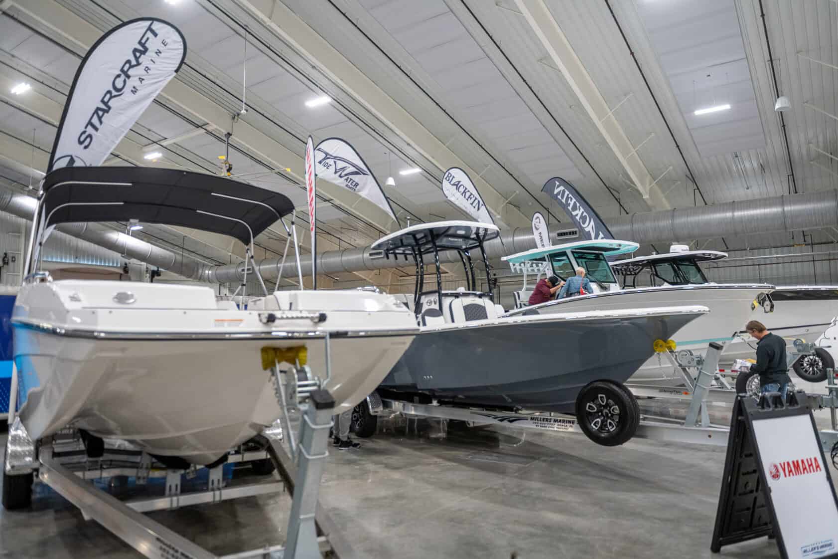 Several boats are displayed indoors at a boat show, with people inspecting them. Brand banners and signs are visible above and around the boats.