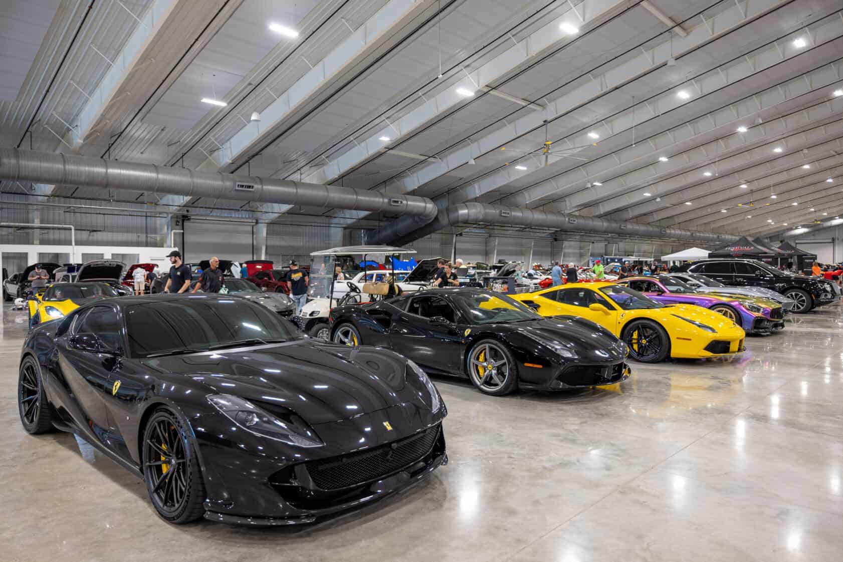 Multiple sports cars, including Ferraris in black, yellow, and red, are displayed in a large indoor exhibition hall with people viewing them.