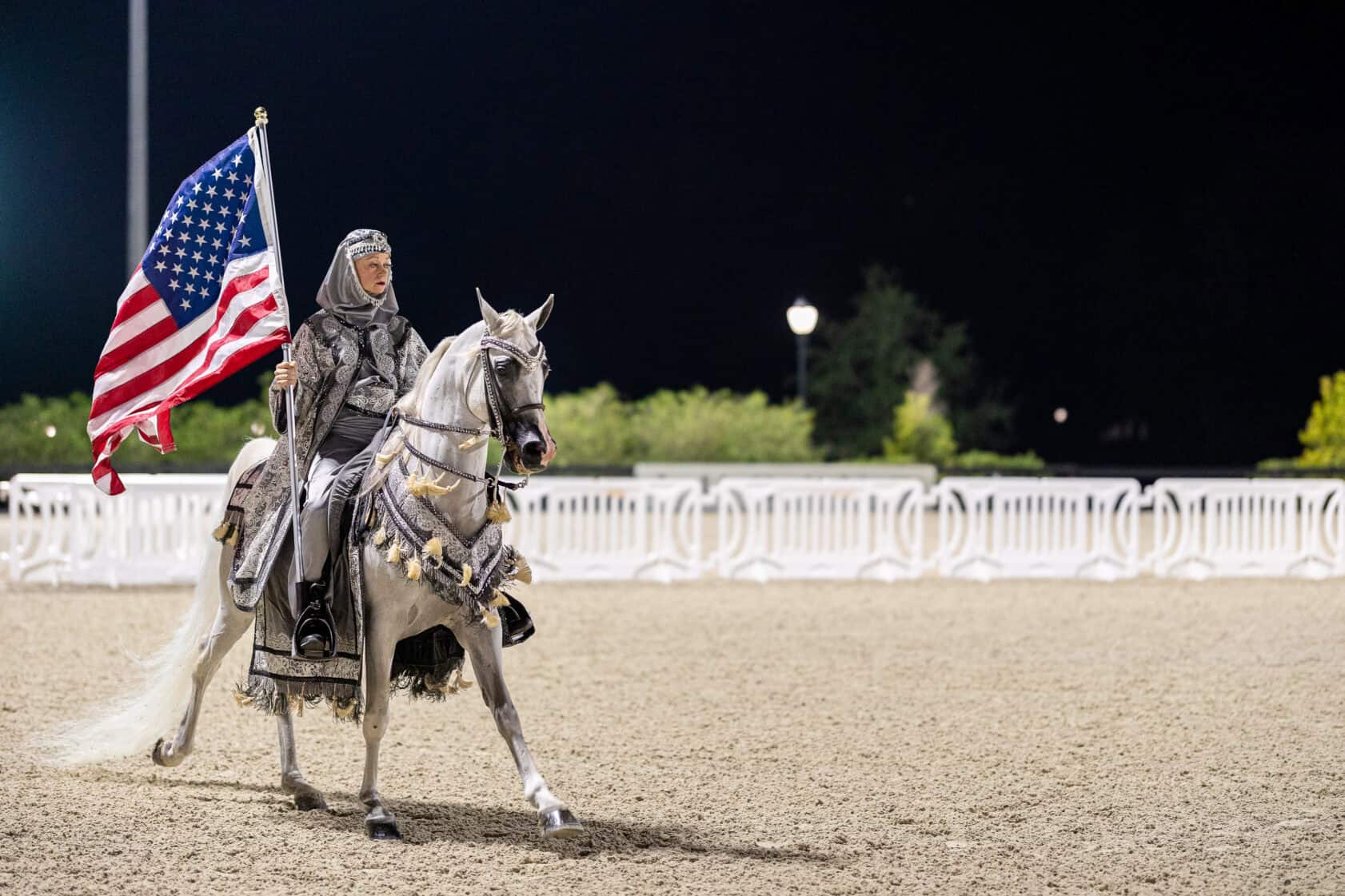 A person in medieval armor rides a white horse and holds an American flag in an outdoor arena at night.