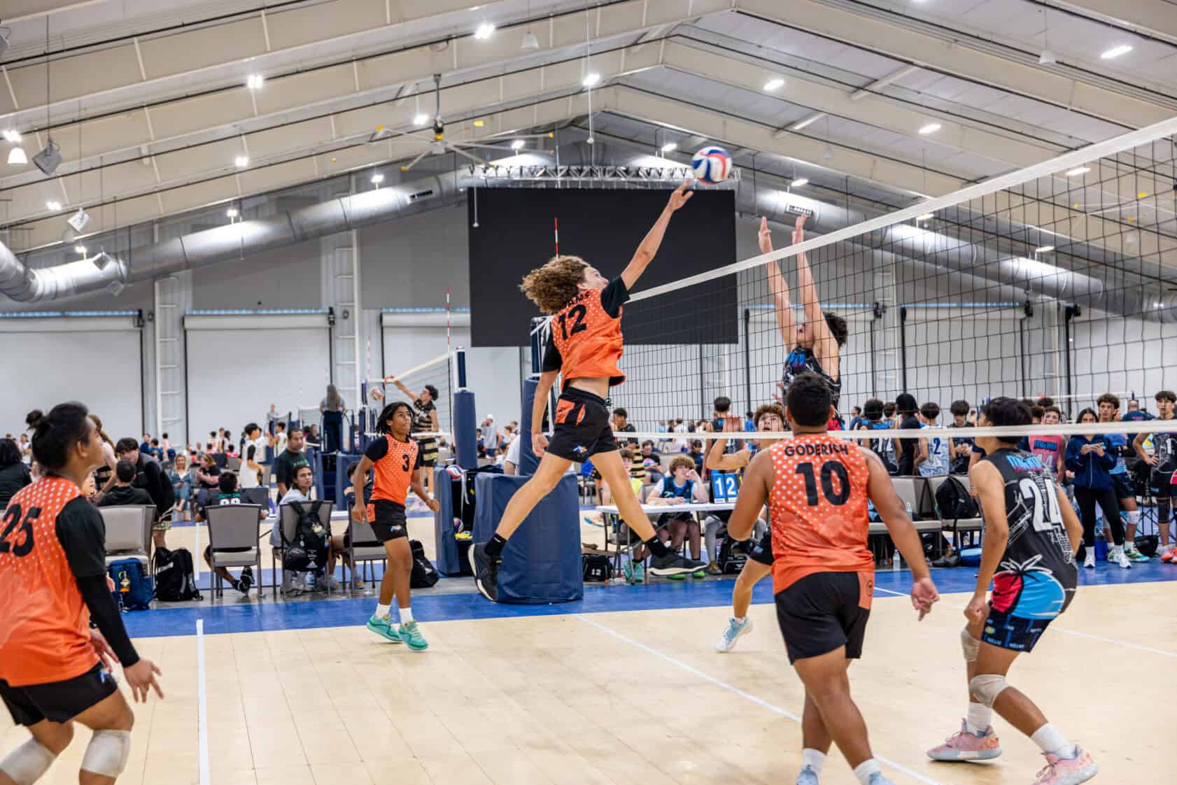 A volleyball player in an orange jersey jumps to spike the ball over the net as opponents and teammates watch during an indoor match.