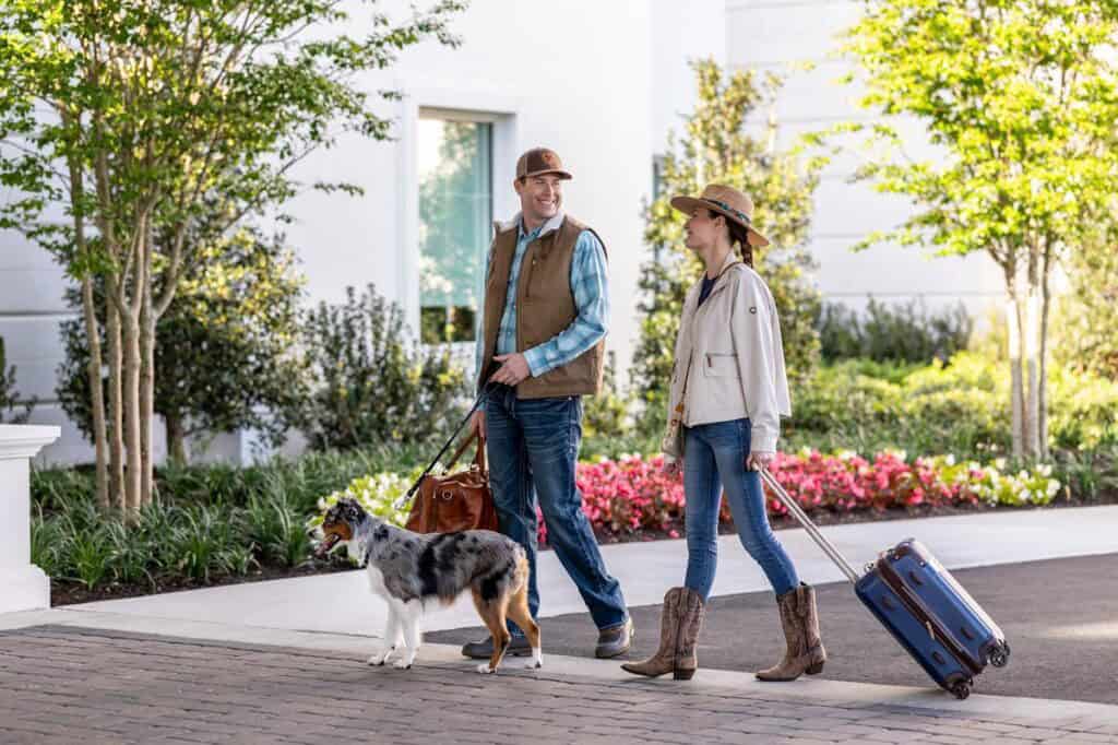 A man and woman walk outside with luggage and a dog on a leash, wearing casual clothes and hats, with greenery and flowers in the background.