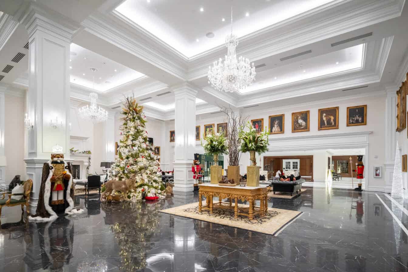 A spacious lobby features marble floors, chandeliers, a decorated Christmas tree, gold table with flowers, and framed portraits on the walls.