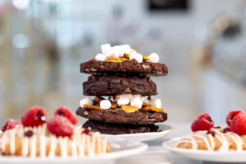 Three chocolate cookies stacked with mini marshmallows and graham crackers, flanked by pastries topped with raspberries and icing on white plates.