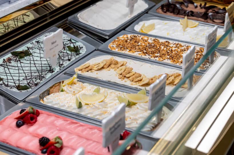 Display case with trays of assorted gelato flavors, including lemon, mint, chocolate, and berry, each decorated with matching toppings.