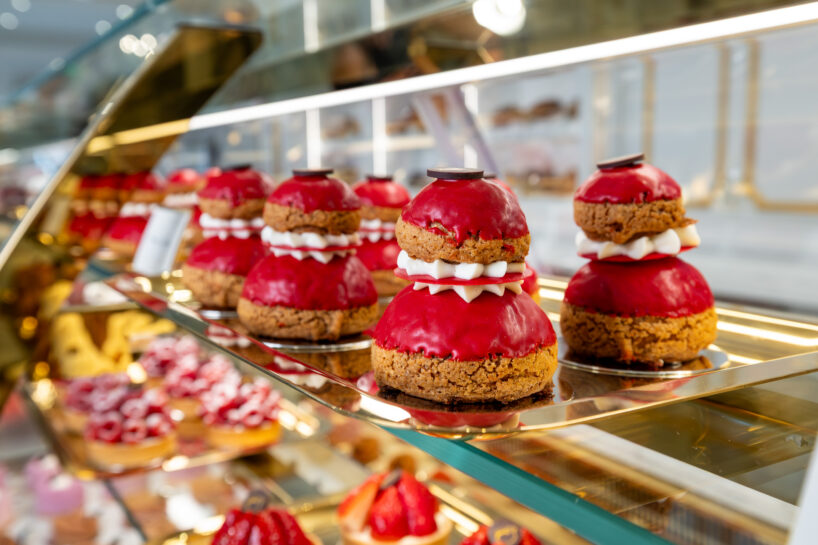 Glass display case with rows of red, cream-filled pastries topped with chocolate discs, alongside other assorted desserts in a bakery.