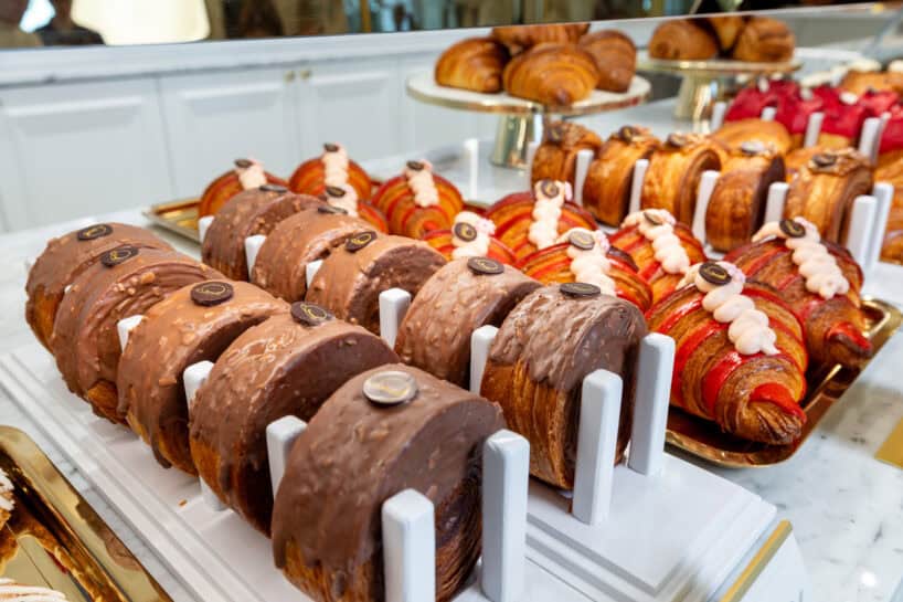 Rows of assorted croissants, including chocolate-dipped and fruit-filled varieties, displayed on trays in a bakery setting.