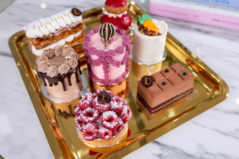 A gold tray with seven assorted, decorative pastries, including fruit tarts, mousse cakes, and layered desserts, arranged on a marble surface.