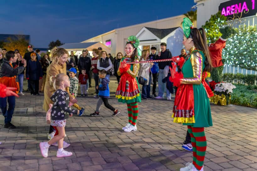 Children and adults watch as two women dressed in colorful costumes hold a limbo stick at an outdoor event in front of a lit building at night.