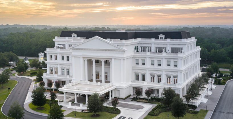 A large, white neoclassical building with columns and multiple stories, surrounded by trees and roads, under a partly cloudy sky at sunrise or sunset.