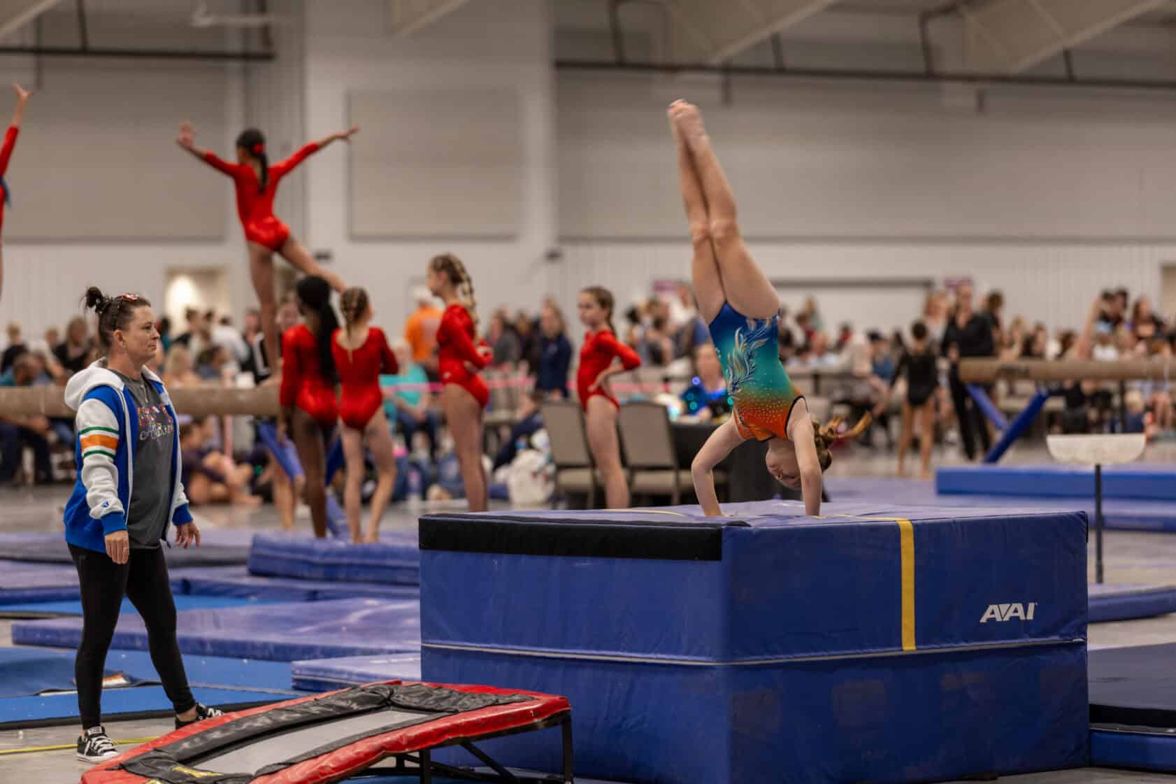A young gymnast does a handstand on a blue mat while others practice nearby; a coach stands watching in a busy gymnasium.