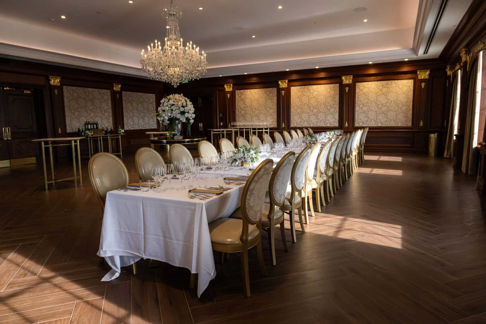 Elegant dining room with a long table set for an event, gold chairs, crystal chandelier, wood floors, and ornate walls.