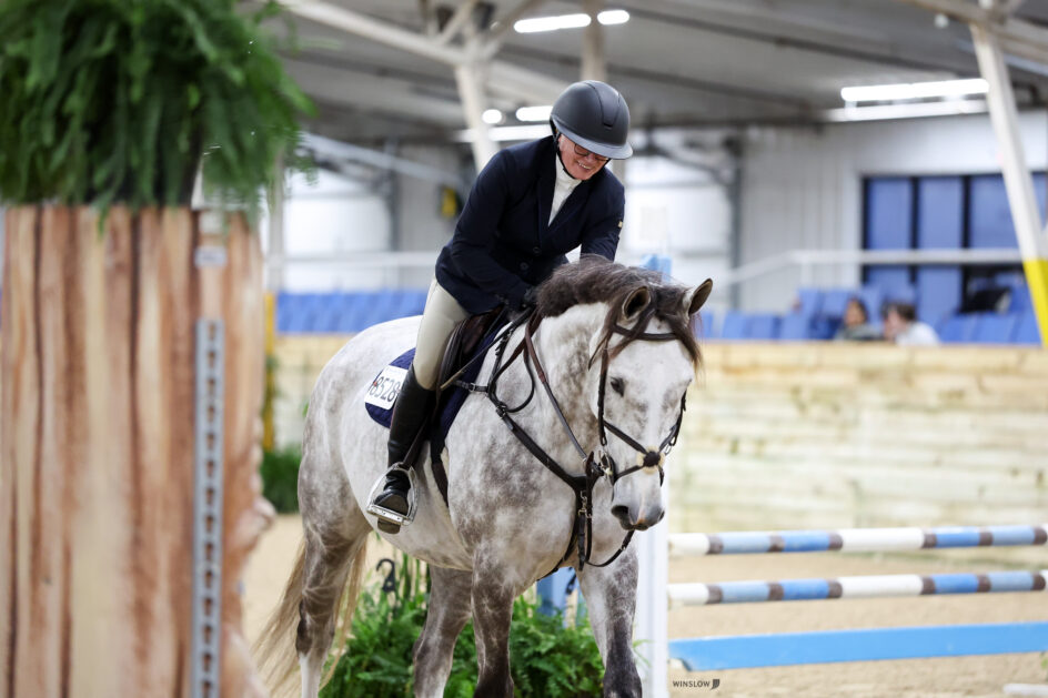 Person in equestrian attire riding a gray horse during an indoor show jumping event, with obstacles and spectators in the background.