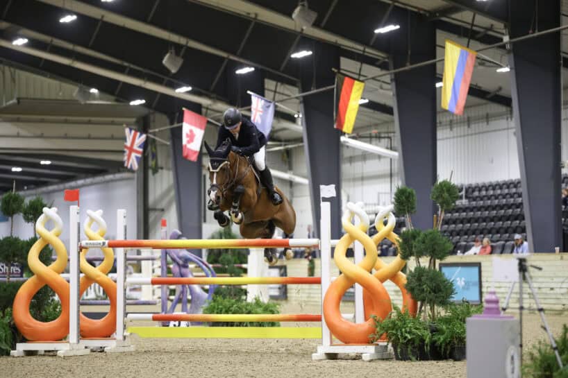 Equestrian athlete in mid-jump on a horse over a colorful obstacle in an indoor arena, with flags hanging above and spectators in the background.