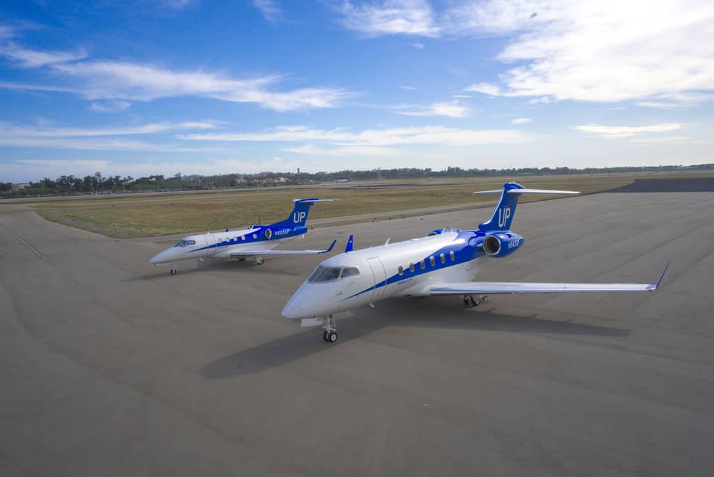 Two blue and white private jets are parked on an empty airport tarmac under a partly cloudy sky.