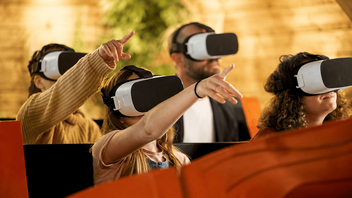 Four people wearing virtual reality headsets sit in a row, reaching forward and pointing, appearing to interact with a virtual environment.